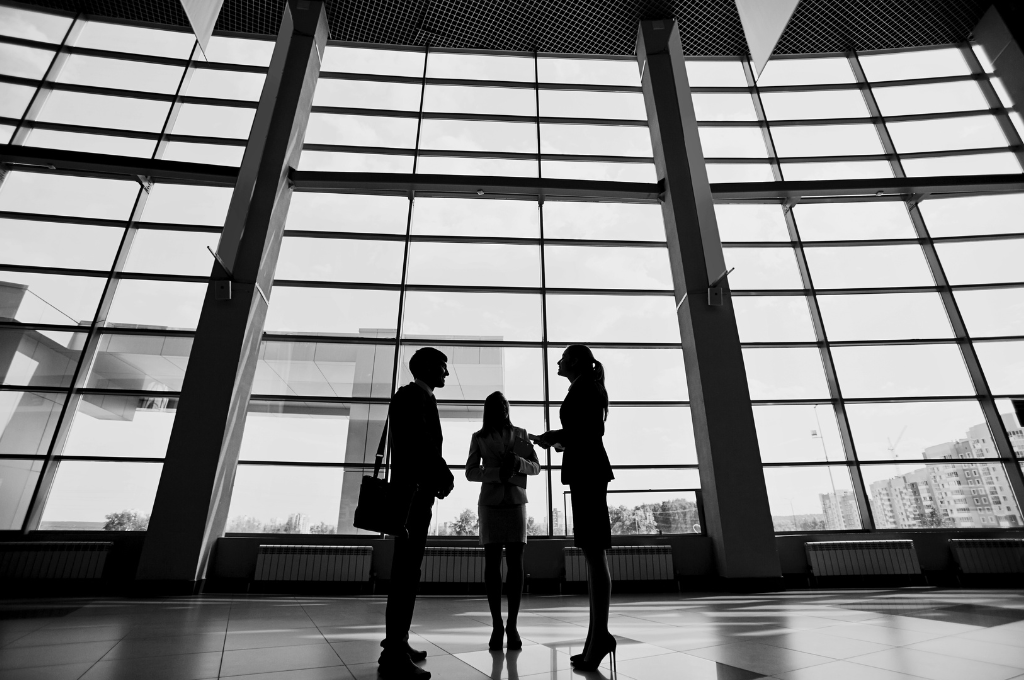 Three people speaking discreetly in a large lobby area with tile and high ceilings. We only see their silhouettes and it's two women and one man. The appear to be representing private intelligence services.