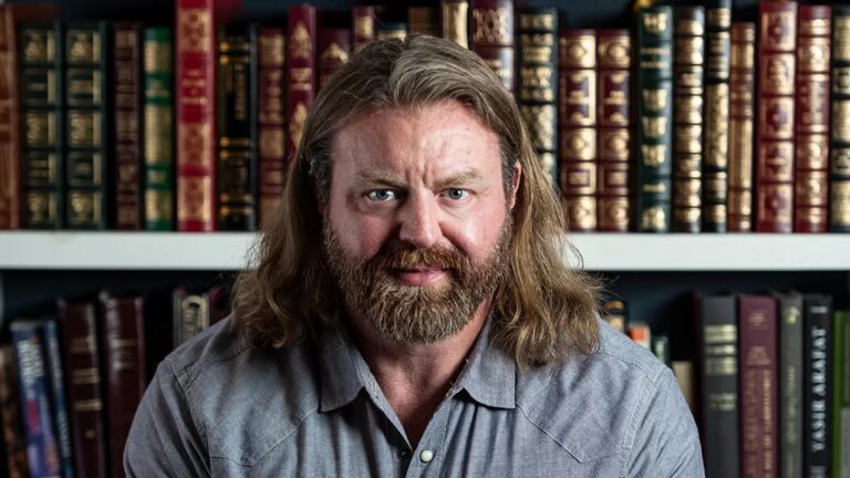 A photo of Bryan Hubbard from Kentucky, He has long hair and a beard, standing in front of a wall library filled with books. Very studious.