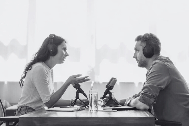 A man and a woman talking on a podcast episode. Looks like they're recording live and in a casual but engaging conversation that they're both enjoying. They're both wearing headphones.